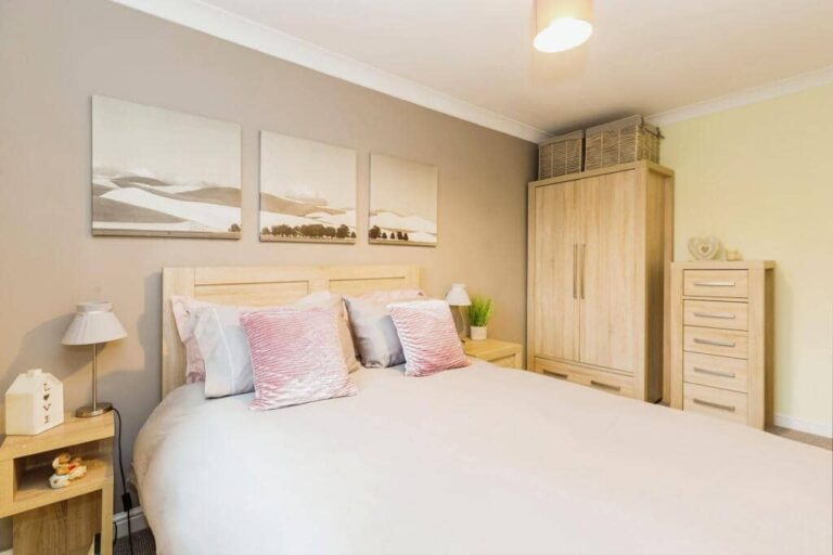 A modern bedroom with a double bed, pink cushions, wooden furniture, and landscape artwork above the headboard.