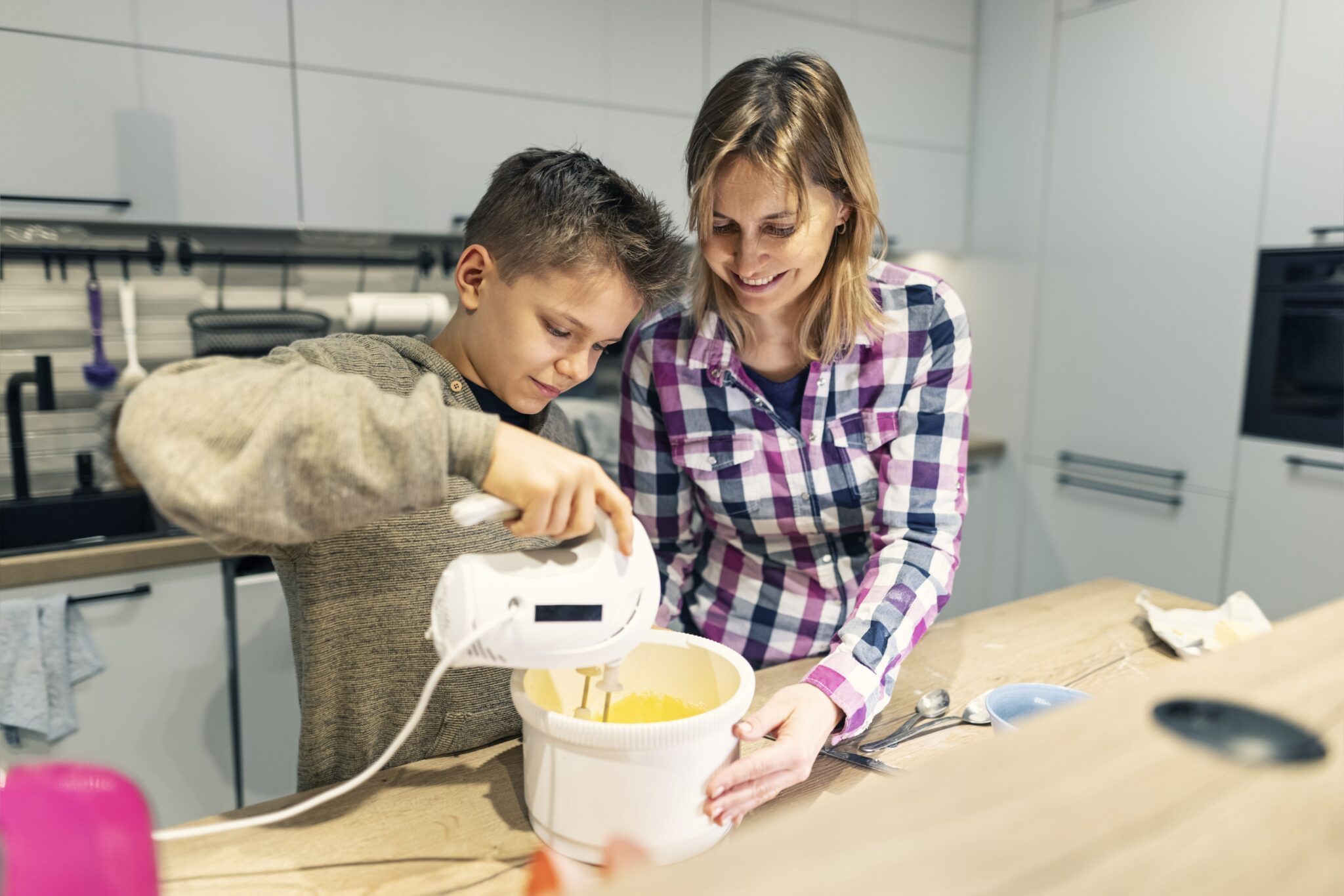 A woman and a child are using an electric mixer to blend ingredients in a kitchen.