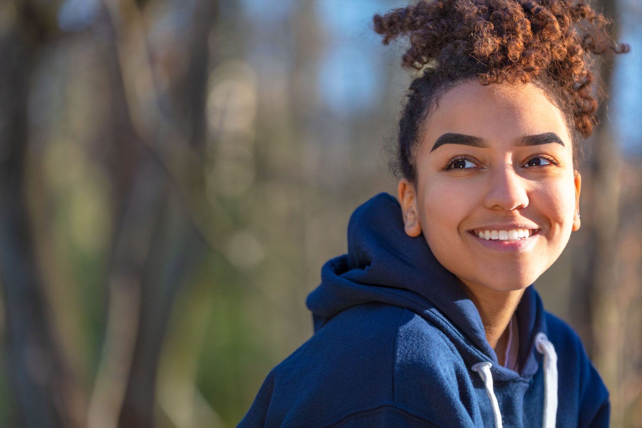 Woman with curly hair smiling, wearing a navy hoodie, outdoors.