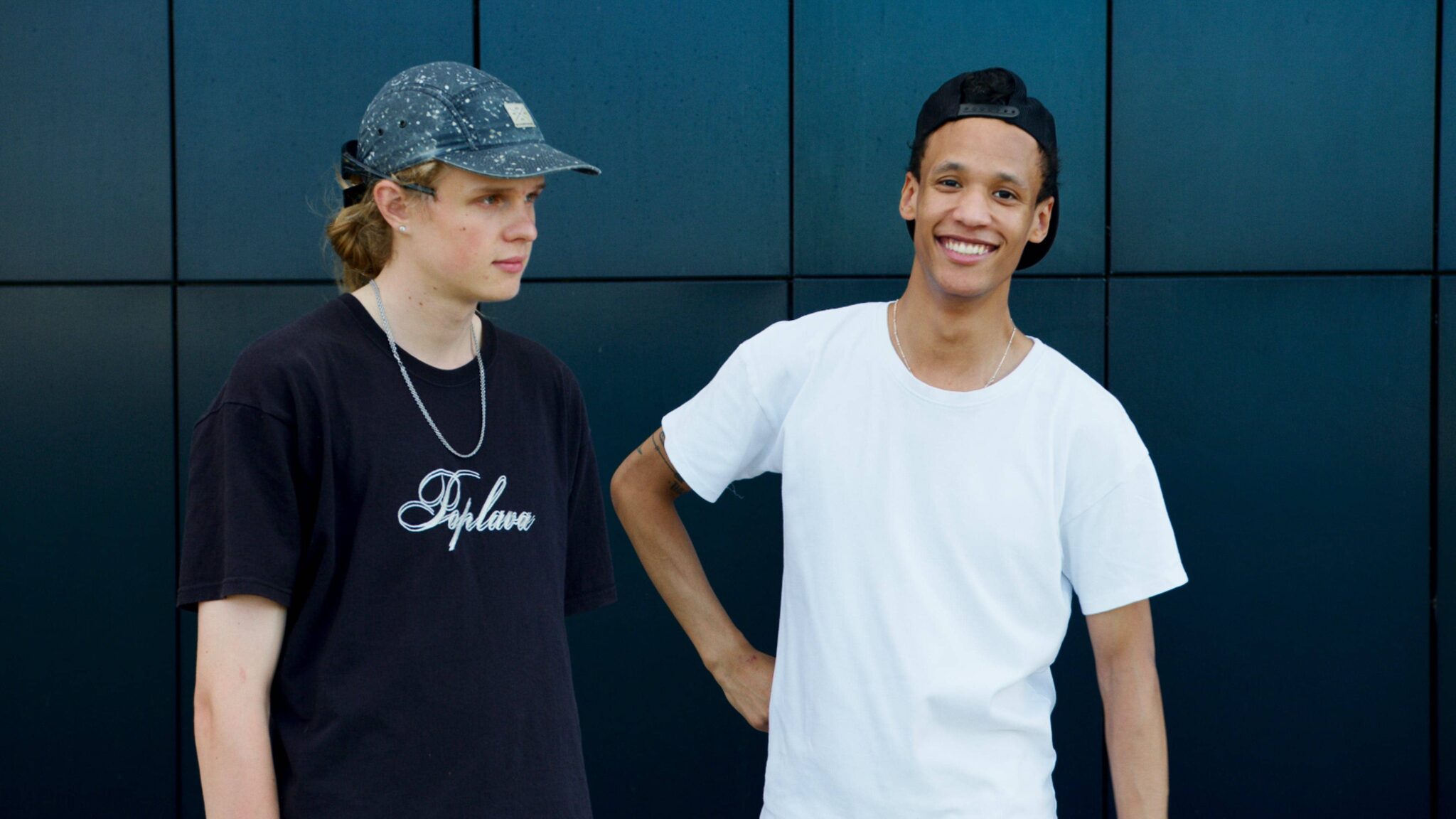 Two young men stand in front of a dark wall. One wears a cap and black shirt, the other a black cap backward and a white shirt, smiling with one hand on his hip.